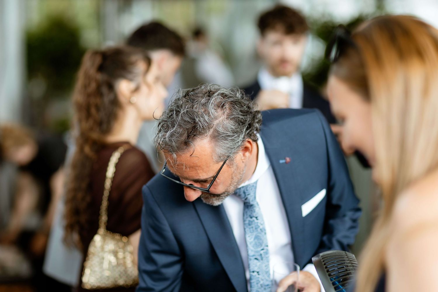 Older man in blue suit looks down at phone among wedding guests outdoors.