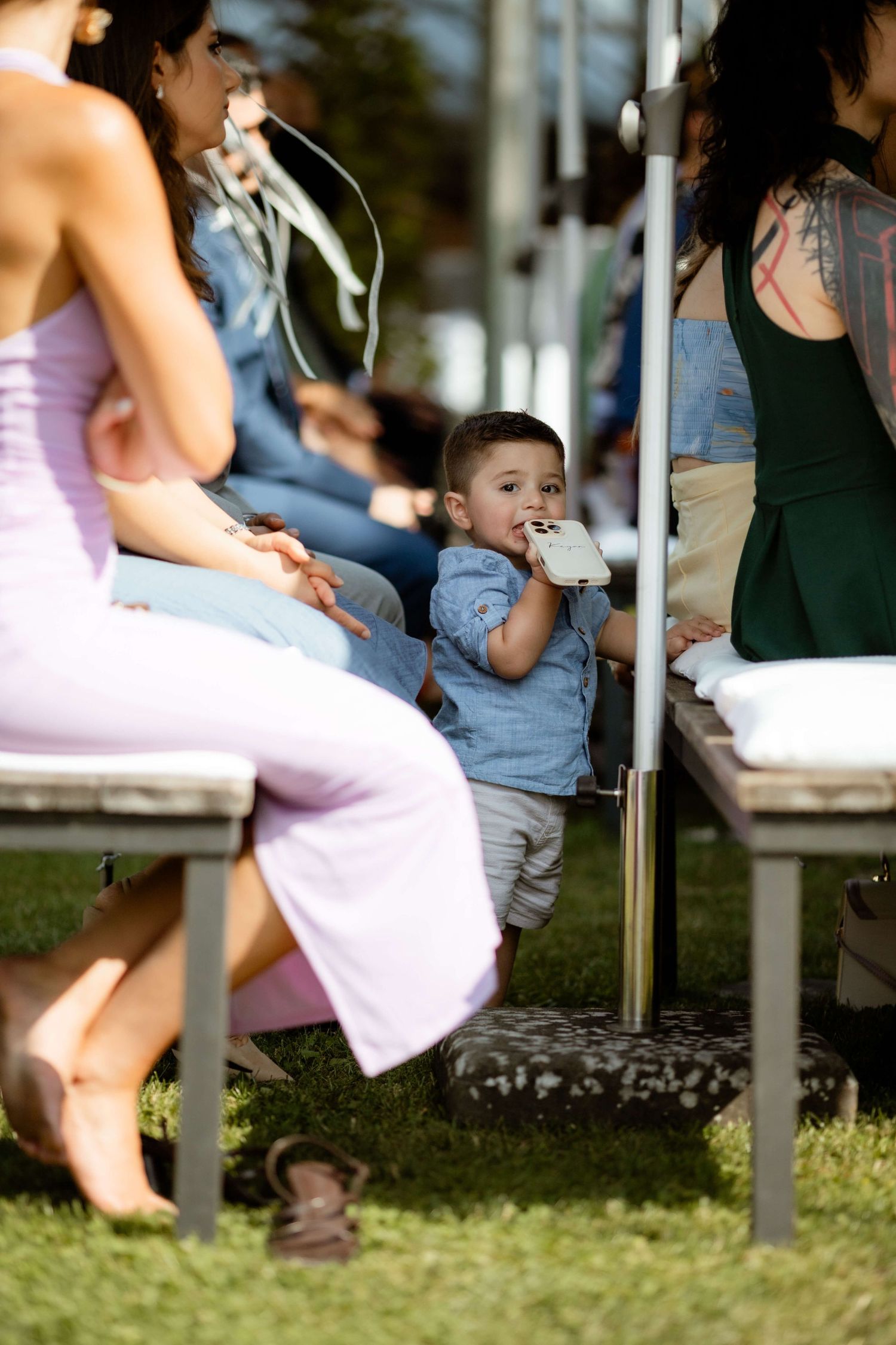 Adorable toddler in blue shirt sits between guests during outdoor wedding ceremony.