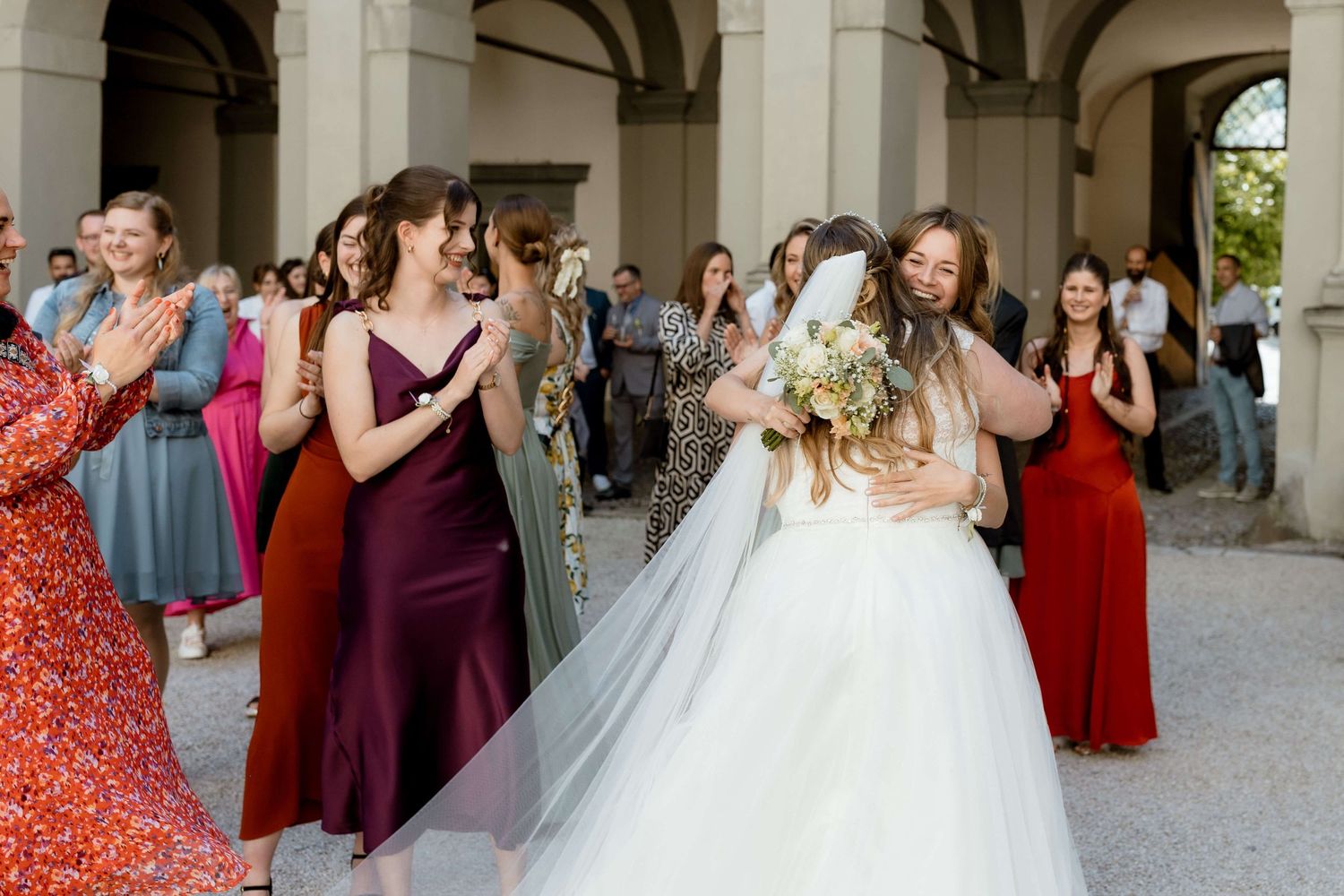 Bride in white gown hugging bridesmaid while wedding guests in colorful dresses applaud outdoors.