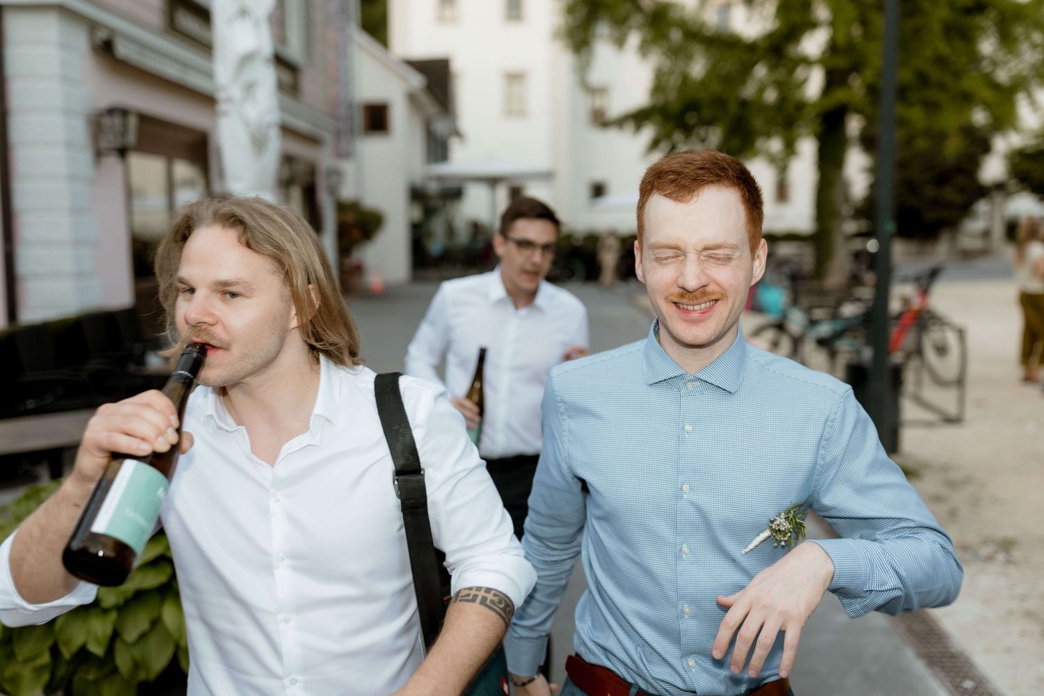 Two smiling men drinking beer bottles walk together outdoors on a city street at a wedding event.
