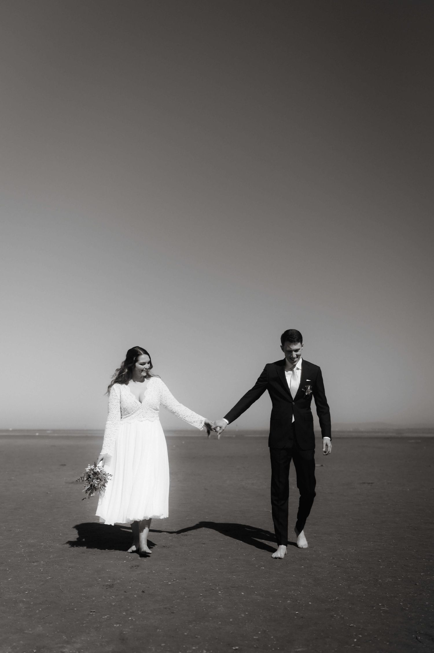 Black and white photo of bride and groom holding hands walking on beach.