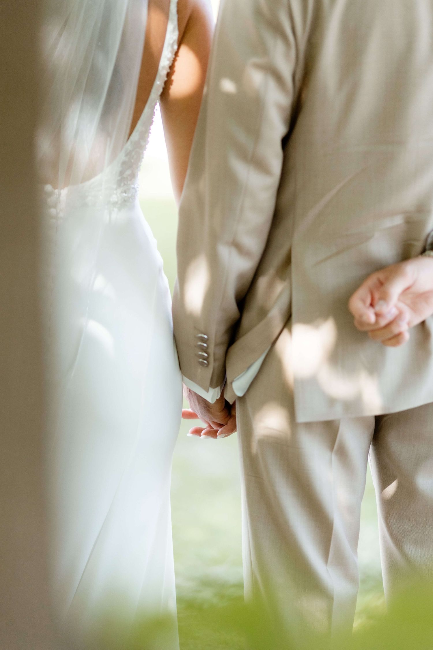 Bride and groom holding hands tenderly, bride in lace gown with veil, groom in tan suit.