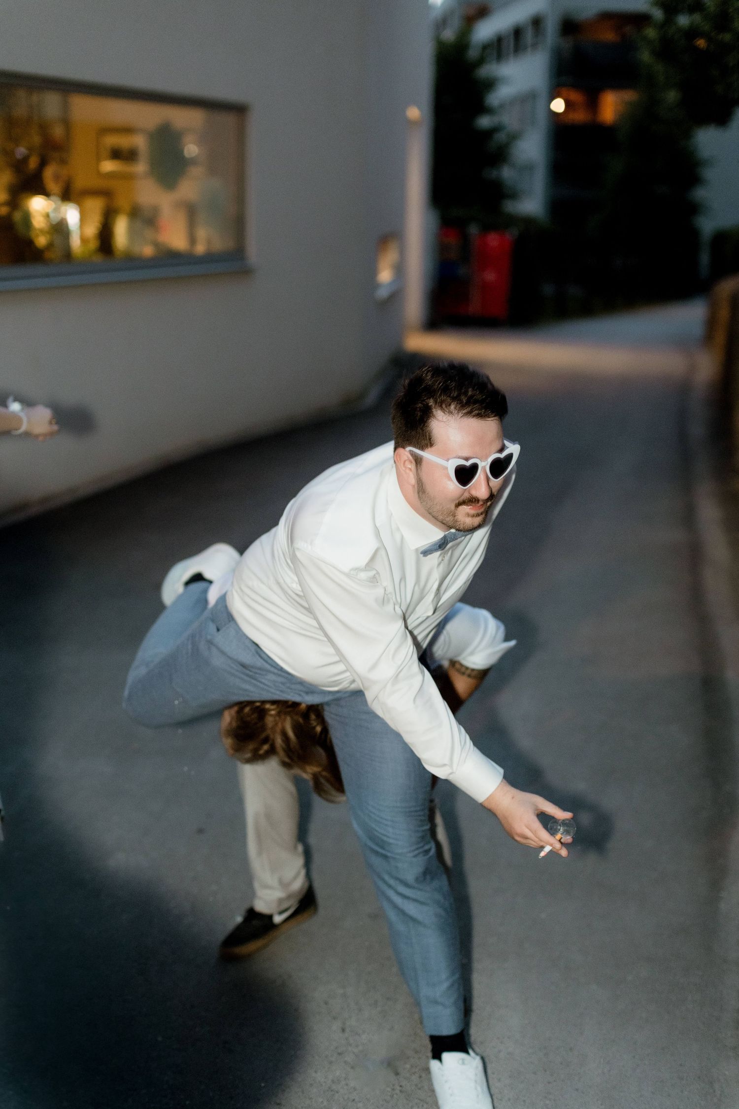 Man in heart sunglasses and white shirt bending down outdoors at a wedding event.