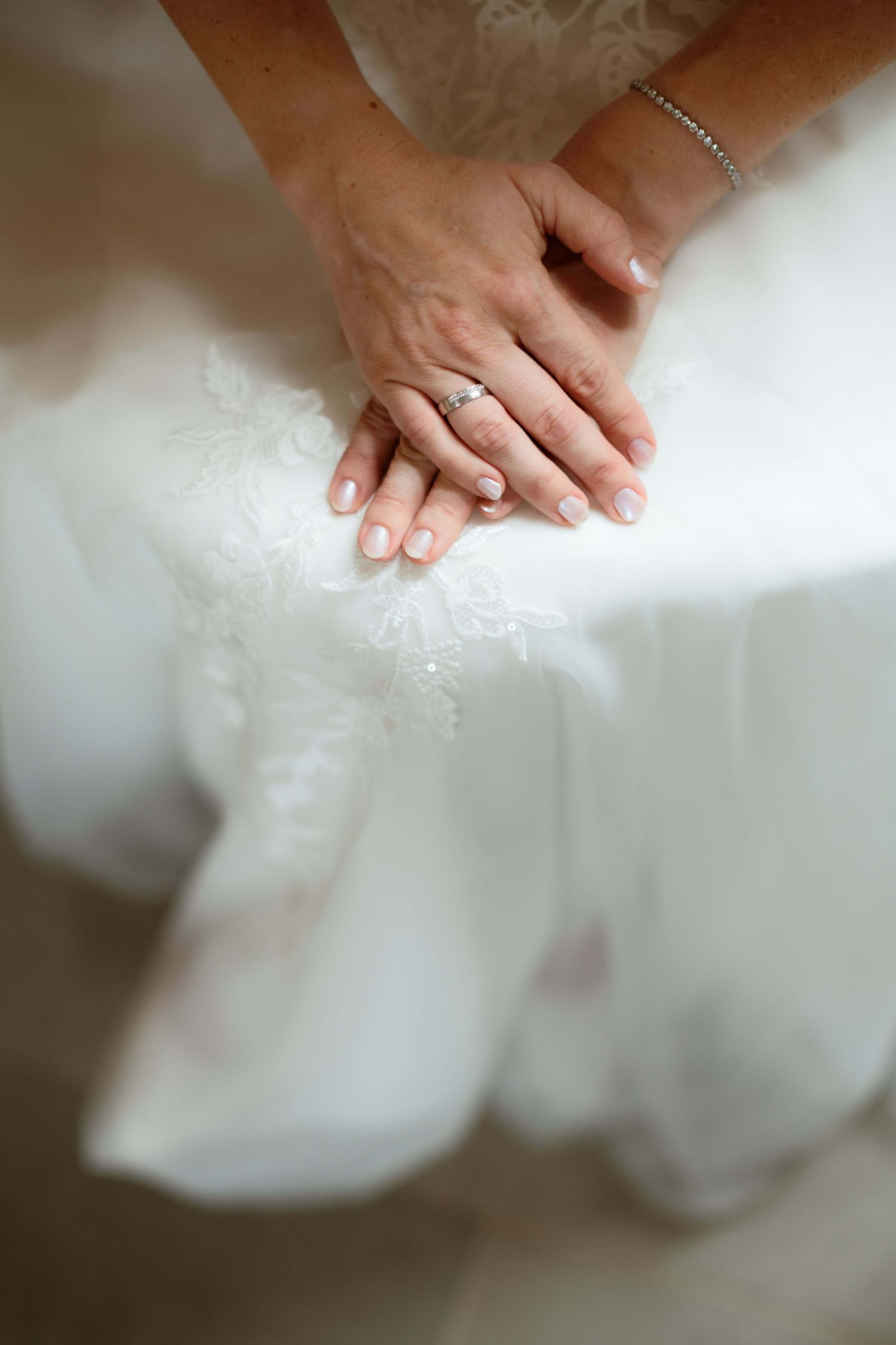 Close-up of bride's hands resting on white lace wedding dress with diamond engagement ring.