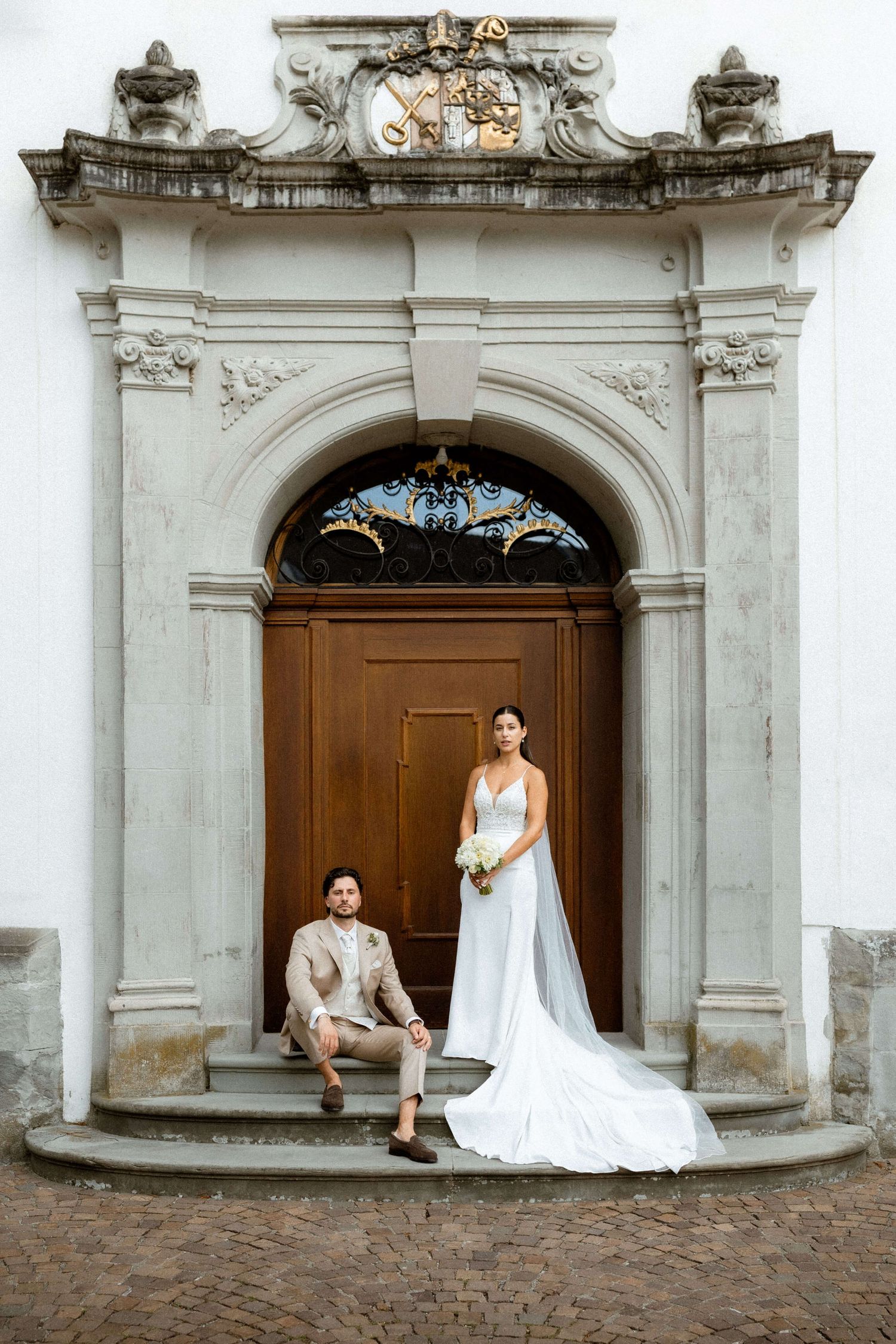 Bride and groom posing before an ornate baroque church doorway on their wedding day.