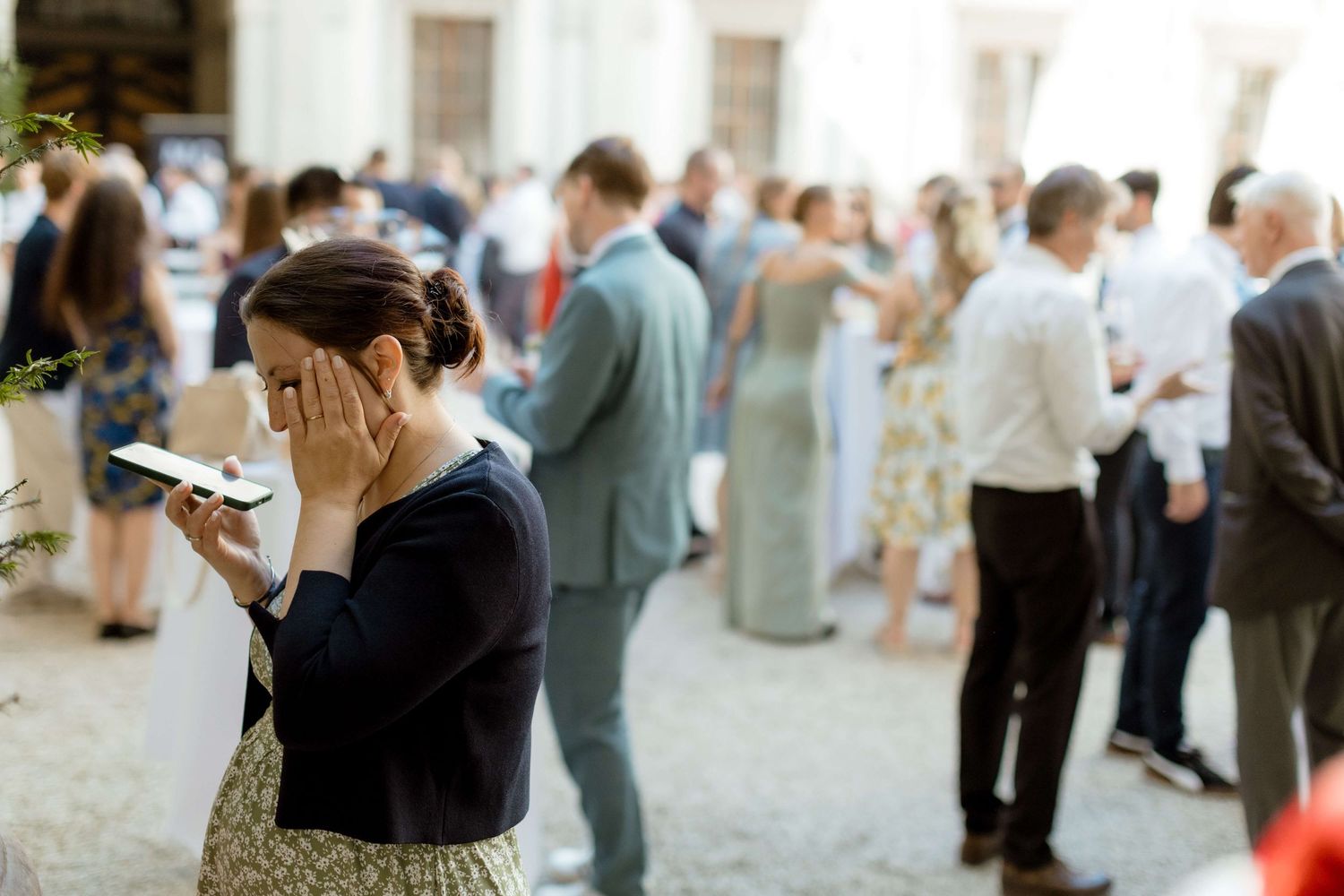 Woman covering face with hand checking phone at outdoor wedding reception with guests mingling.