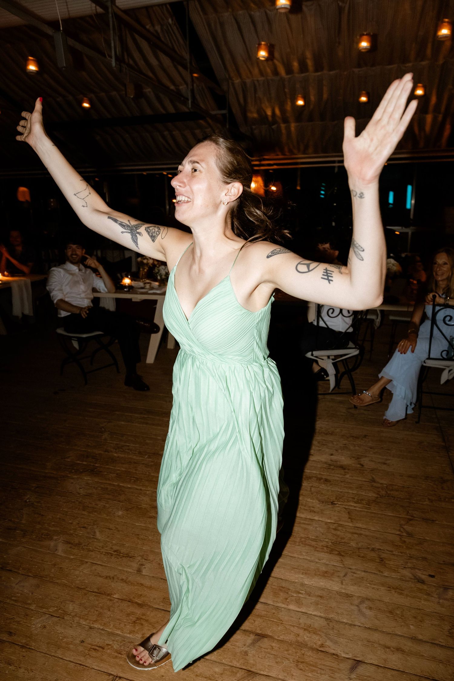 Tattooed woman in mint green dress dancing with arms raised at wedding reception.