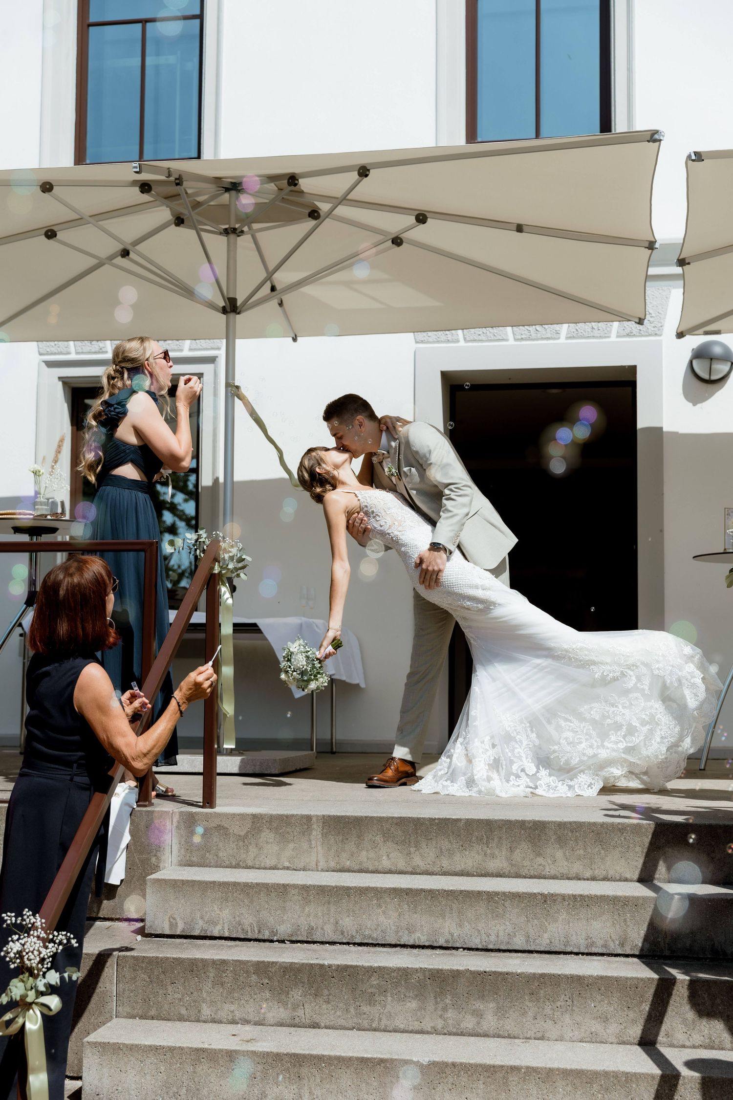 Groom in grey suit dipping bride in white dress for a romantic kiss on sunlit venue steps.
