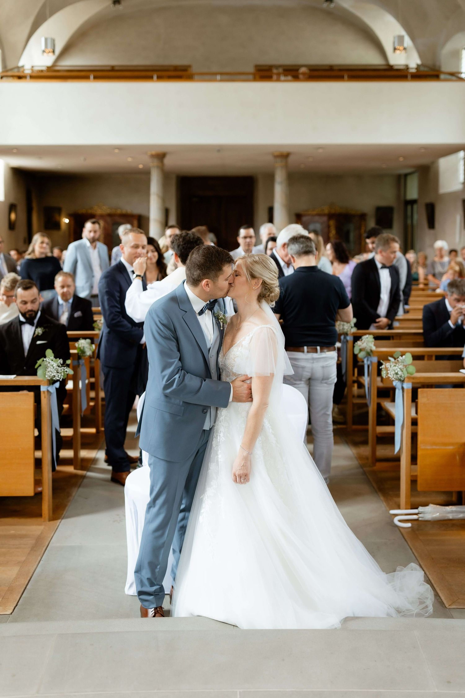 Bride and groom share first kiss at altar during church wedding ceremony with guests watching.