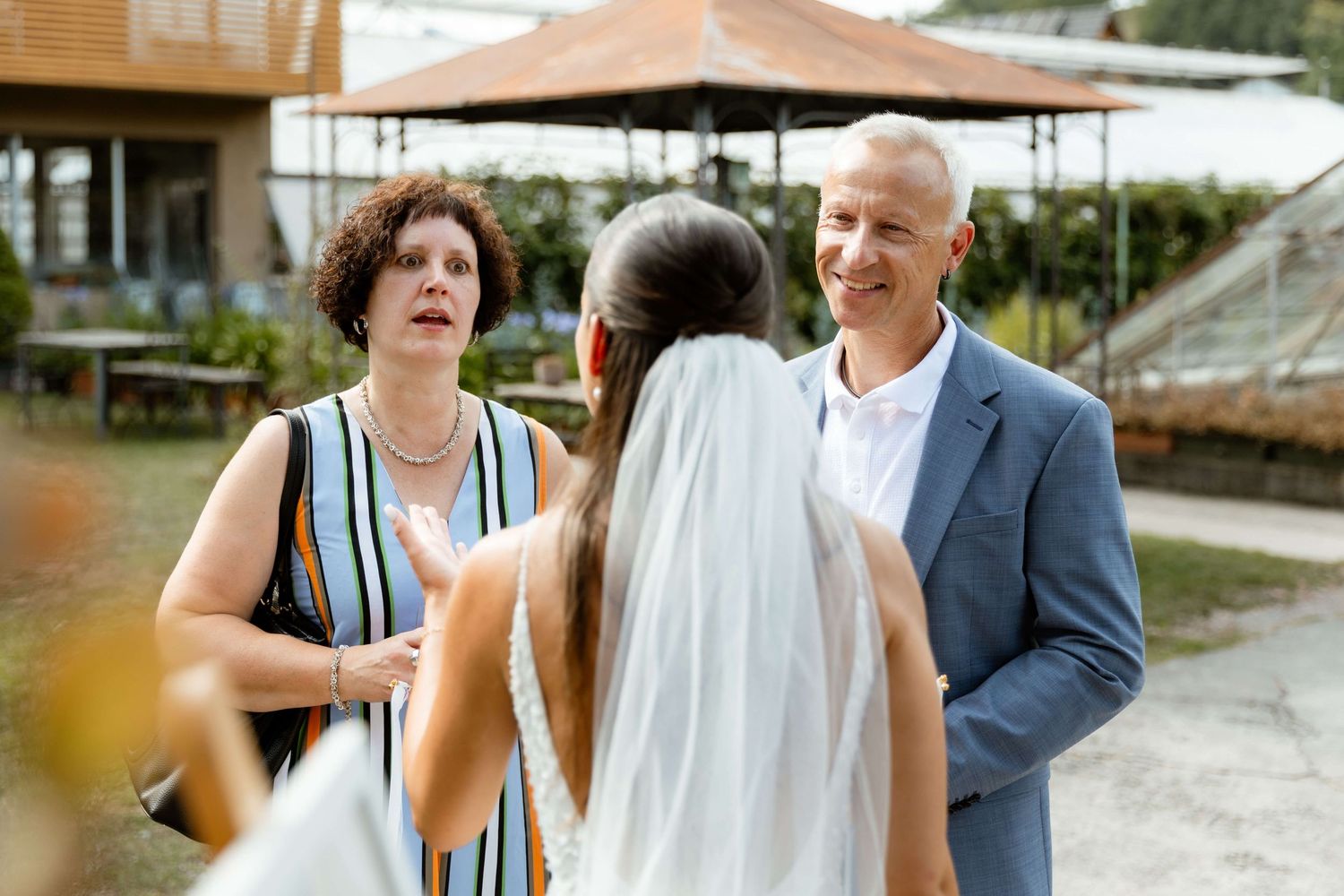 Bride in white veil speaks with older couple at outdoor wedding reception venue.