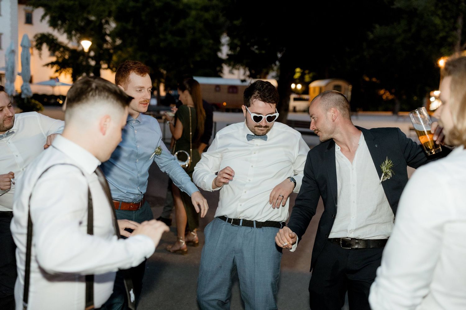 Group of men in formal attire dancing and celebrating outdoors at a wedding reception.
