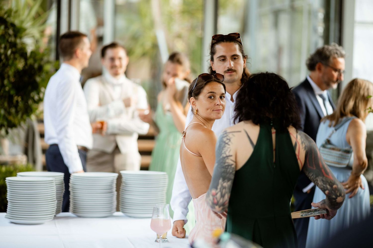 A tattooed bride in a strapless gown glances back at guests near a white tablecloth buffet at a wedding reception.