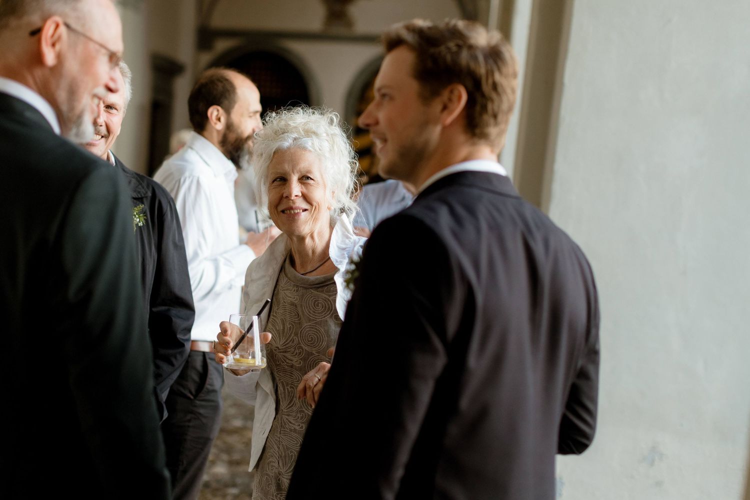 Elderly woman smiling warmly while conversing with guests in elegant archway wedding venue.