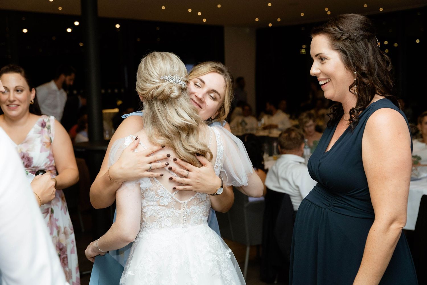 A bride in a lace wedding dress receives a warm hug from a guest at her wedding reception.