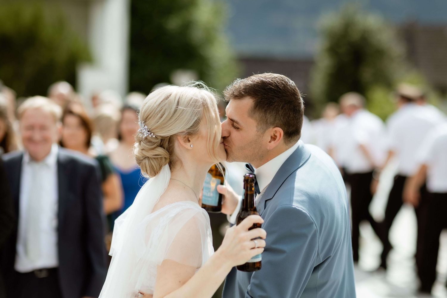 Bride and groom sharing a kiss while holding beer bottles at outdoor wedding reception.