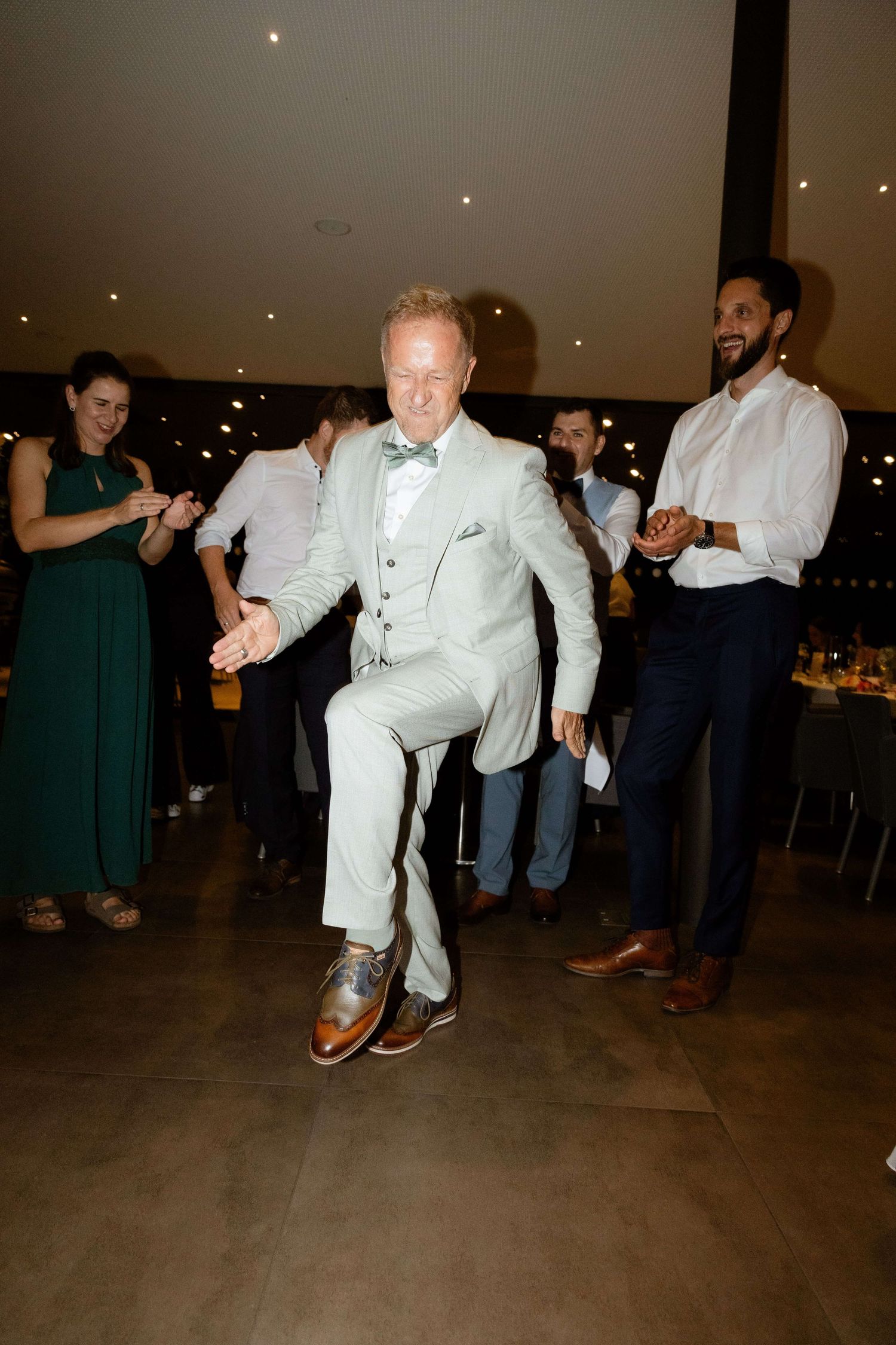 A man in a light gray suit dances energetically on a wedding reception dance floor with guests watching.