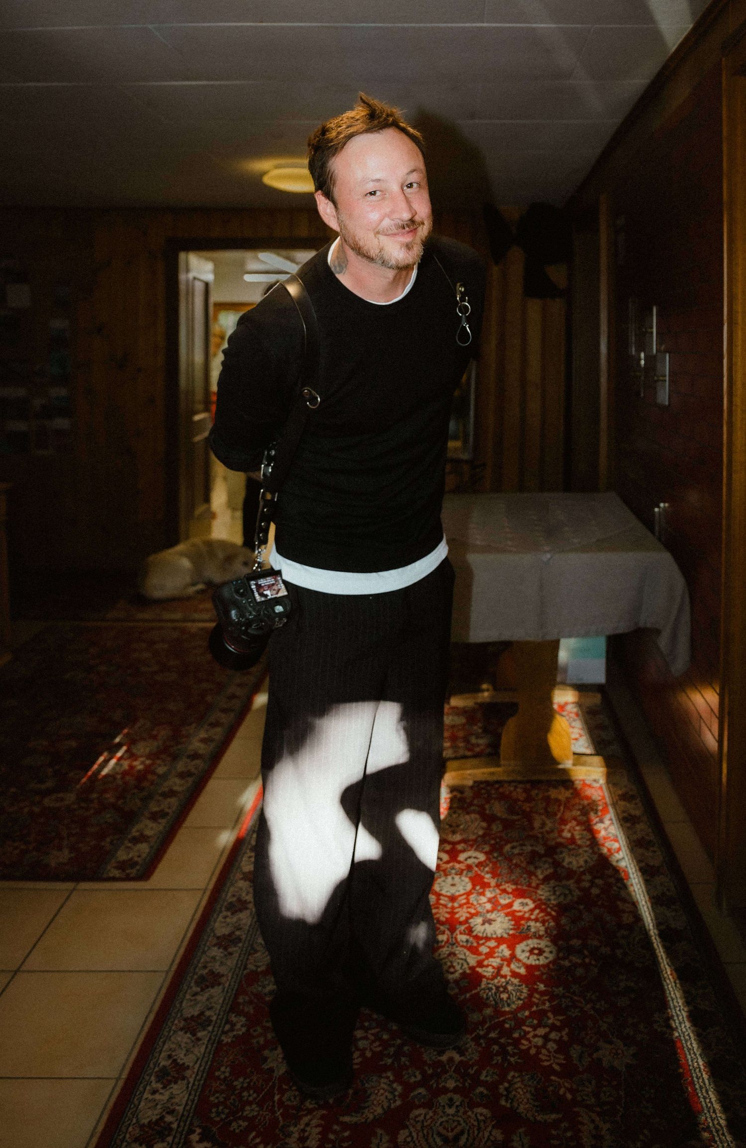 Man in black outfit carrying camera walking down hallway with patterned rug.