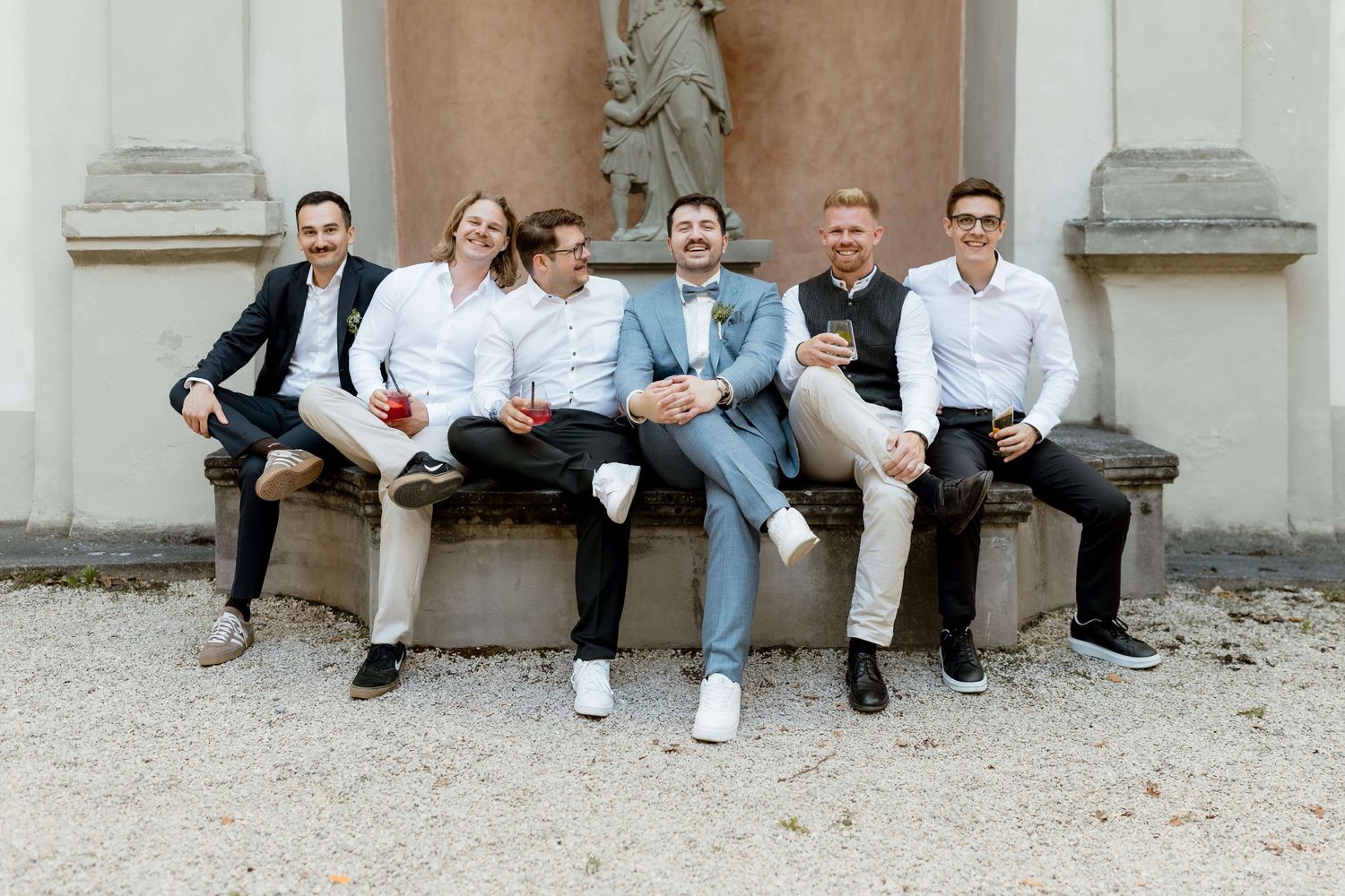 Six groomsmen smiling and sitting on a stone bench outdoors at a sunny wedding celebration.