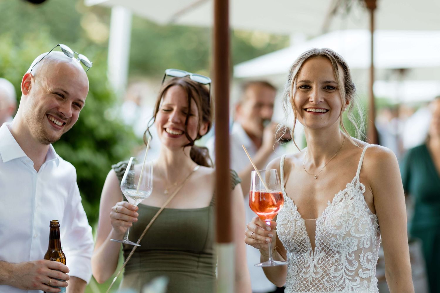 Bride in lace gown holds rosé wine while smiling with guests at outdoor wedding reception.