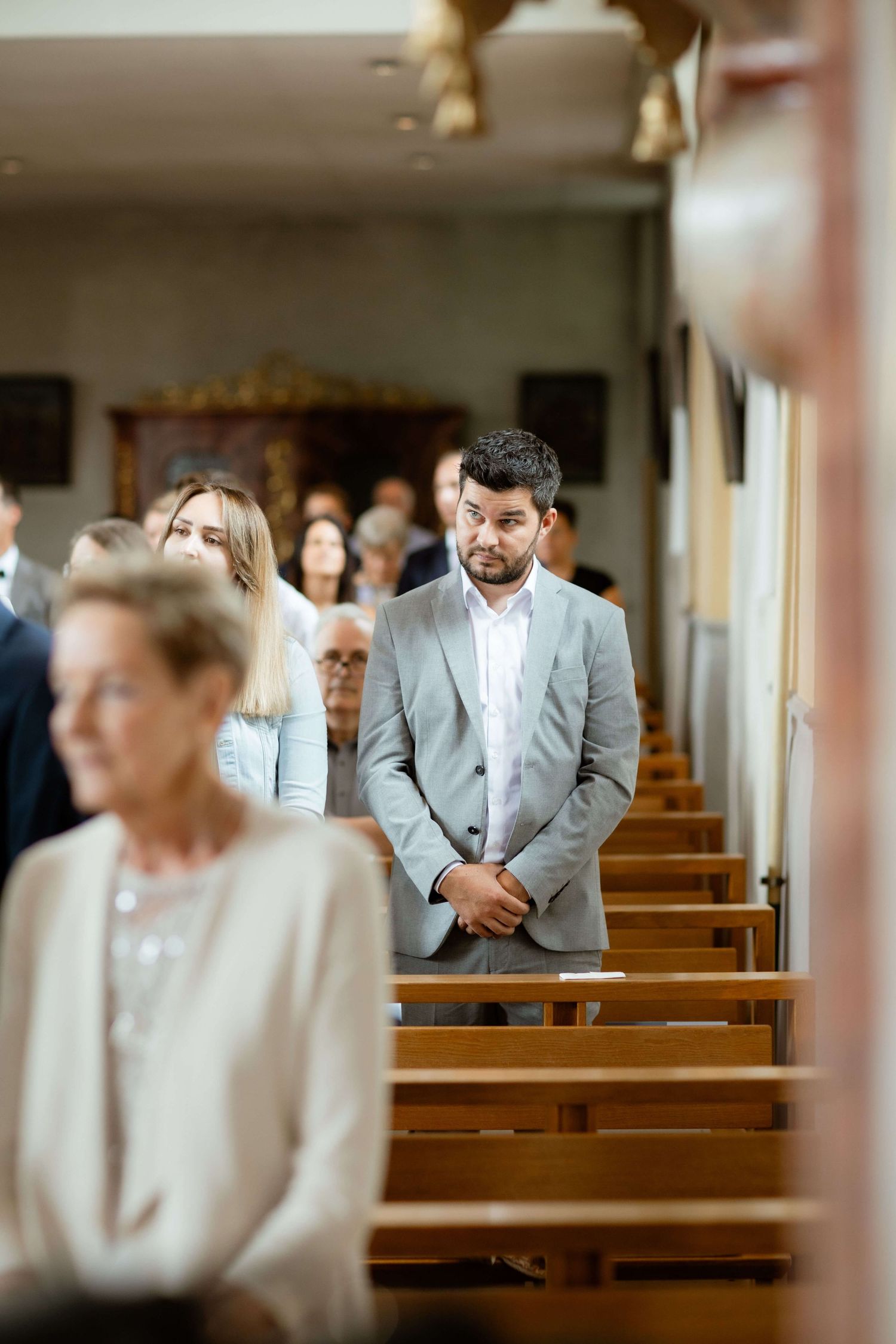 Groom in grey suit standing attentively in church aisle during wedding ceremony.