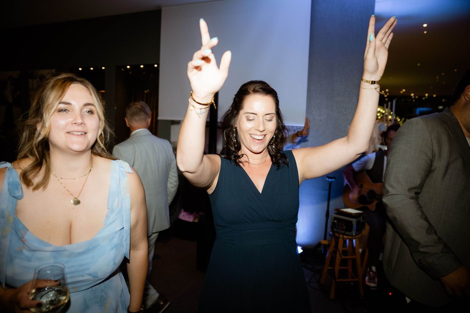 Two women dance joyfully at a wedding reception, one in a navy dress with arms raised high.
