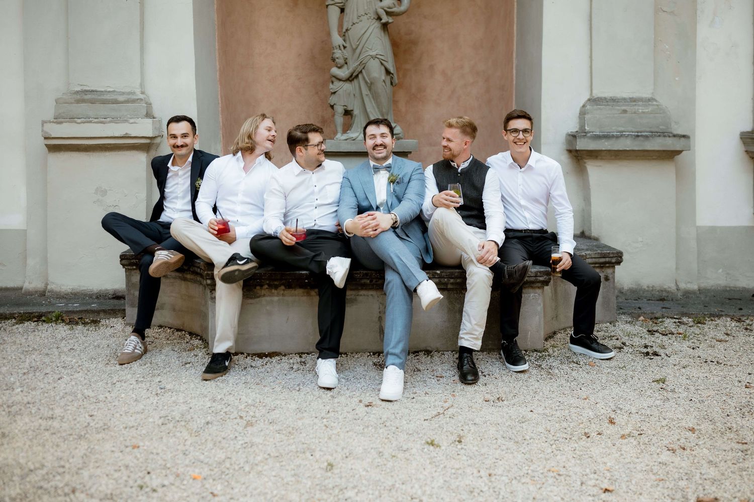 Group of six friends laughing and sitting on a stone bench in front of a statue at a wedding.