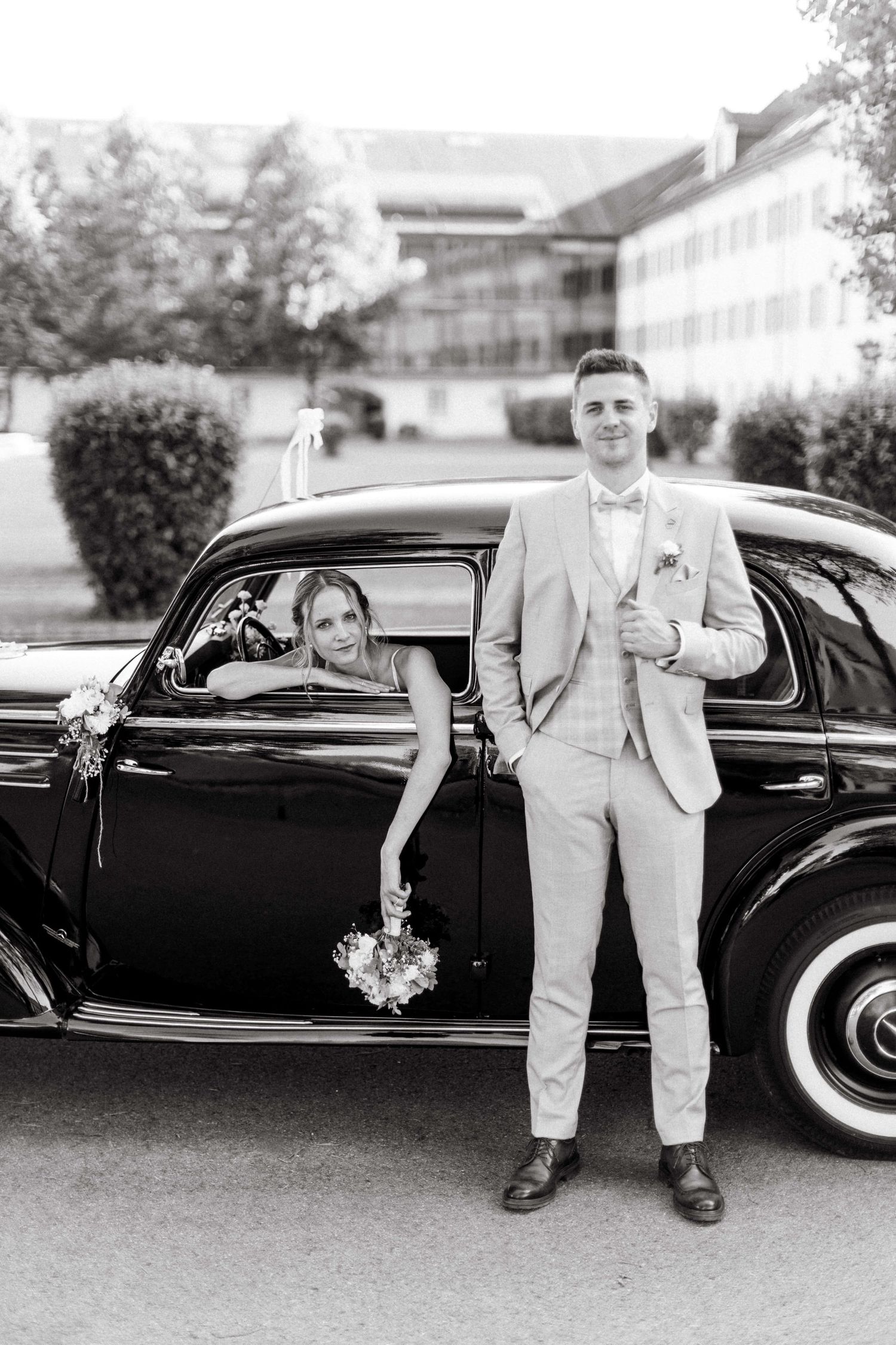 Groom in light suit standing by vintage black car as bride leans out window holding bouquet, black and white.