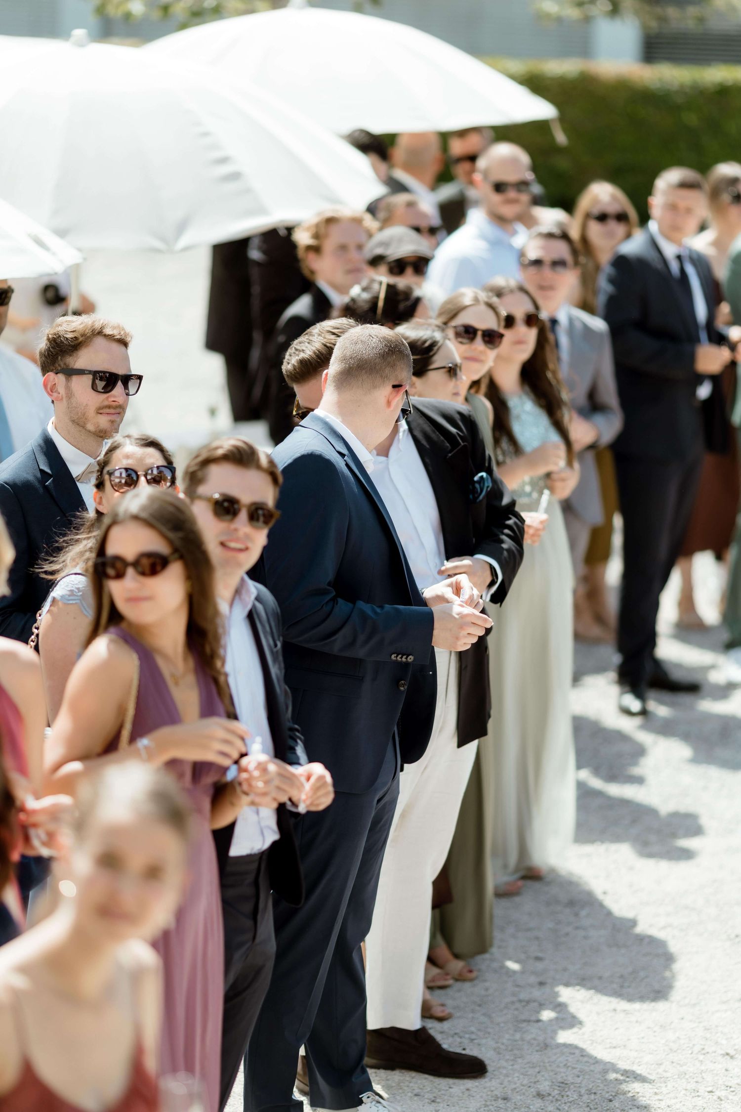 Wedding guests in formal attire stand in line outdoors under white umbrellas on a sunny day.