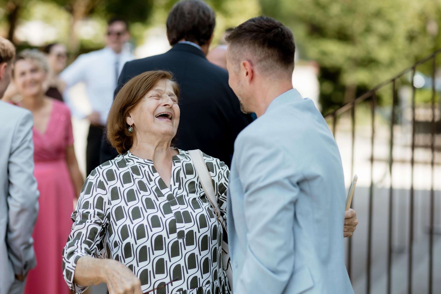 Older woman laughs joyfully while conversing with groom in light gray suit at outdoor wedding celebration.