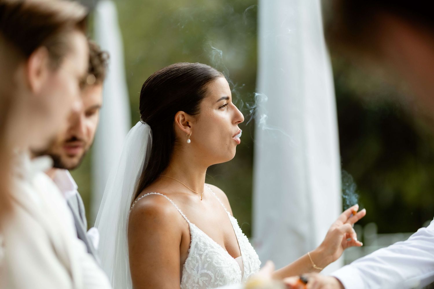 A bride in a white lace gown and veil exhales cigarette smoke while seated outdoors at her wedding ceremony.