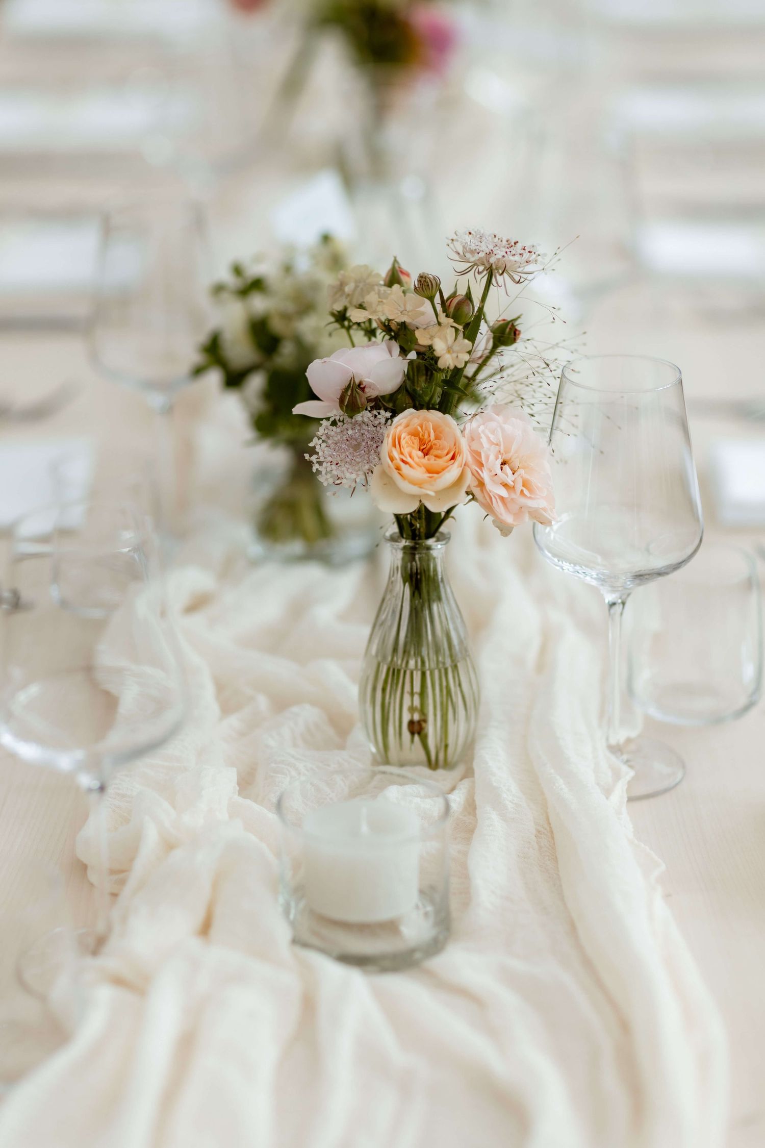 Delicate peach roses in glass vase with candle on white linen wedding table.