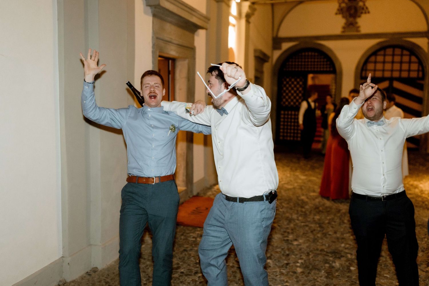Three men celebrate enthusiastically indoors at a wedding reception, raising arms and cheering in a historic venue.
