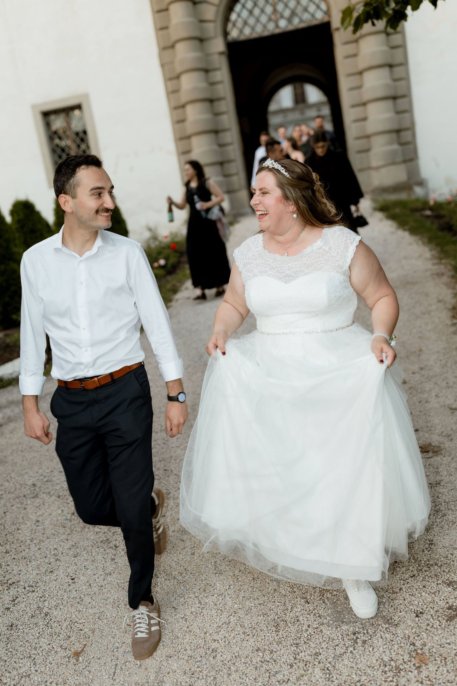 Smiling bride in white tulle dress and groom in white shirt walk joyfully along a gravel path outside a historic building.