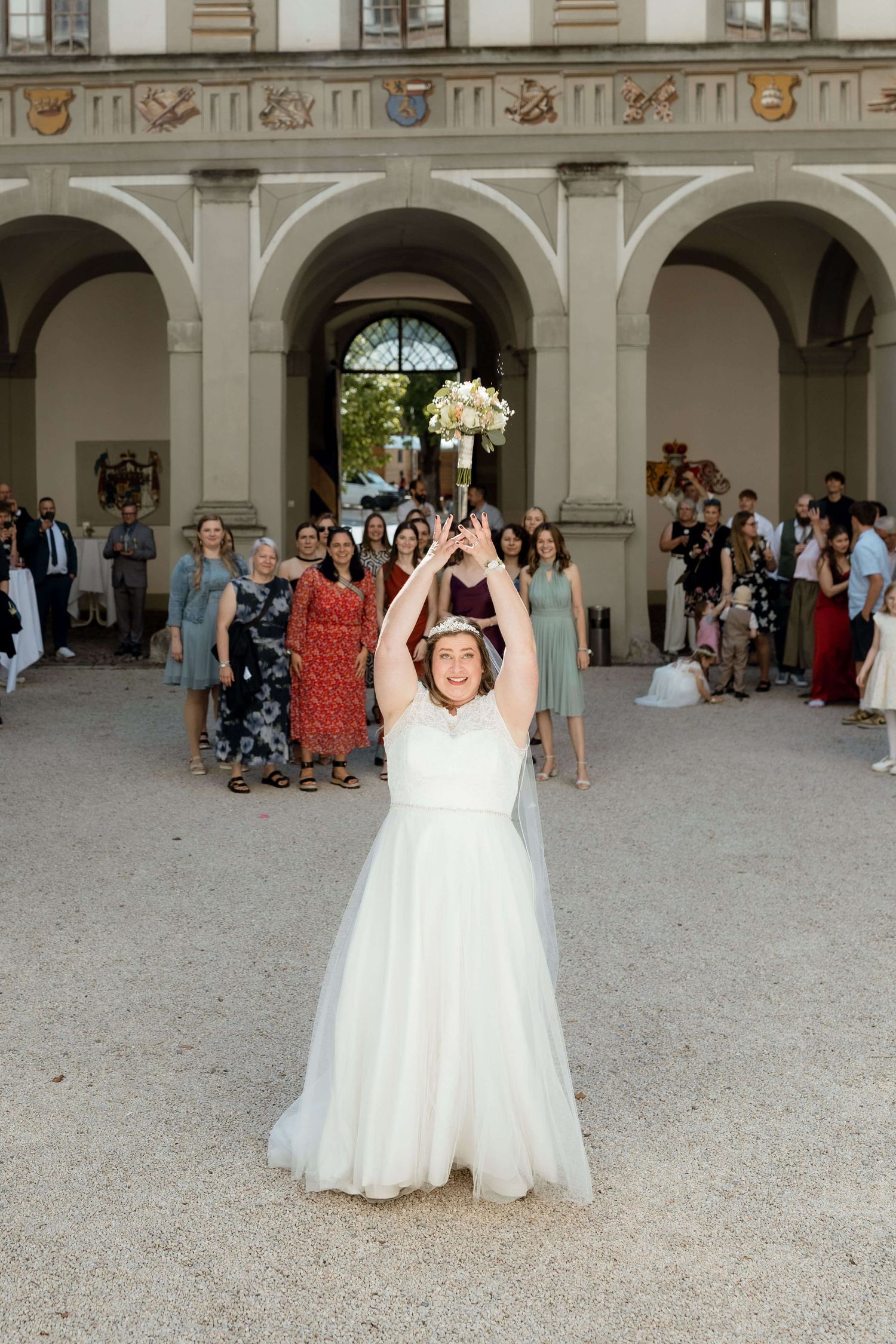 Bride in white tulle gown tossing bouquet to guests at outdoor wedding venue with arches.