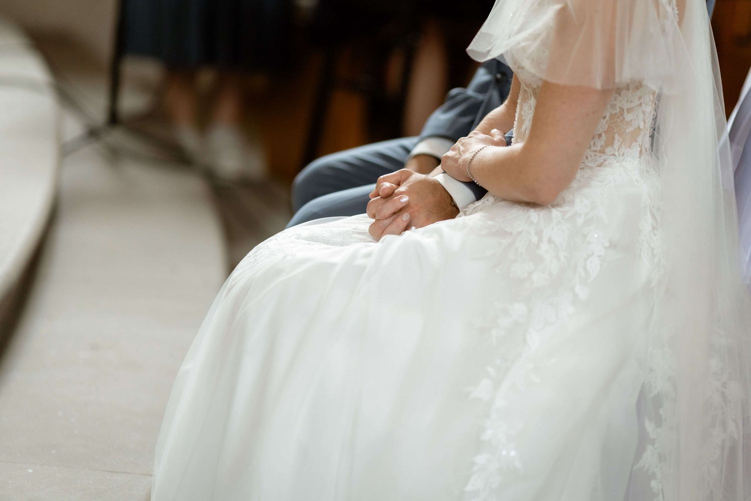 Bride in white lace gown with clasped hands resting on her lap during ceremony.