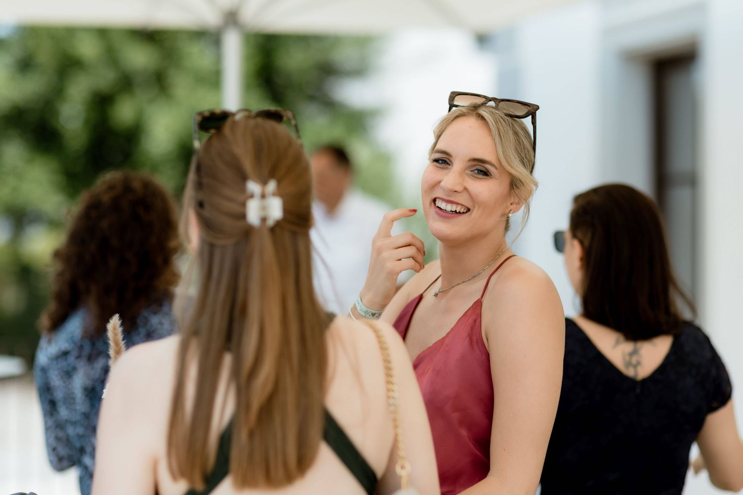 Smiling blonde woman in pink dress holds green apple while chatting with guests at outdoor wedding reception.