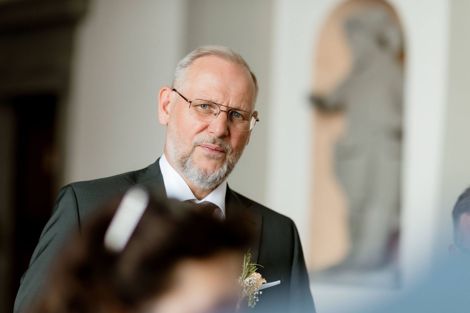 Distinguished older man with glasses and boutonniere looking serious at an elegant wedding ceremony.