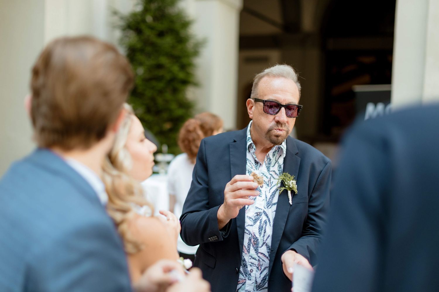 Older man in a floral shirt and blazer with sunglasses enjoying a bite at an outdoor wedding event.