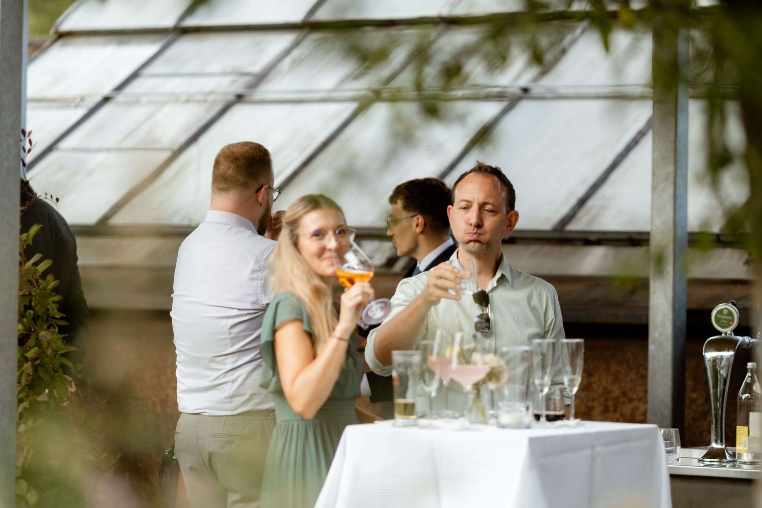 Guests enjoy drinks at a white-clothed table in a greenhouse venue, with a woman sipping an orange cocktail.