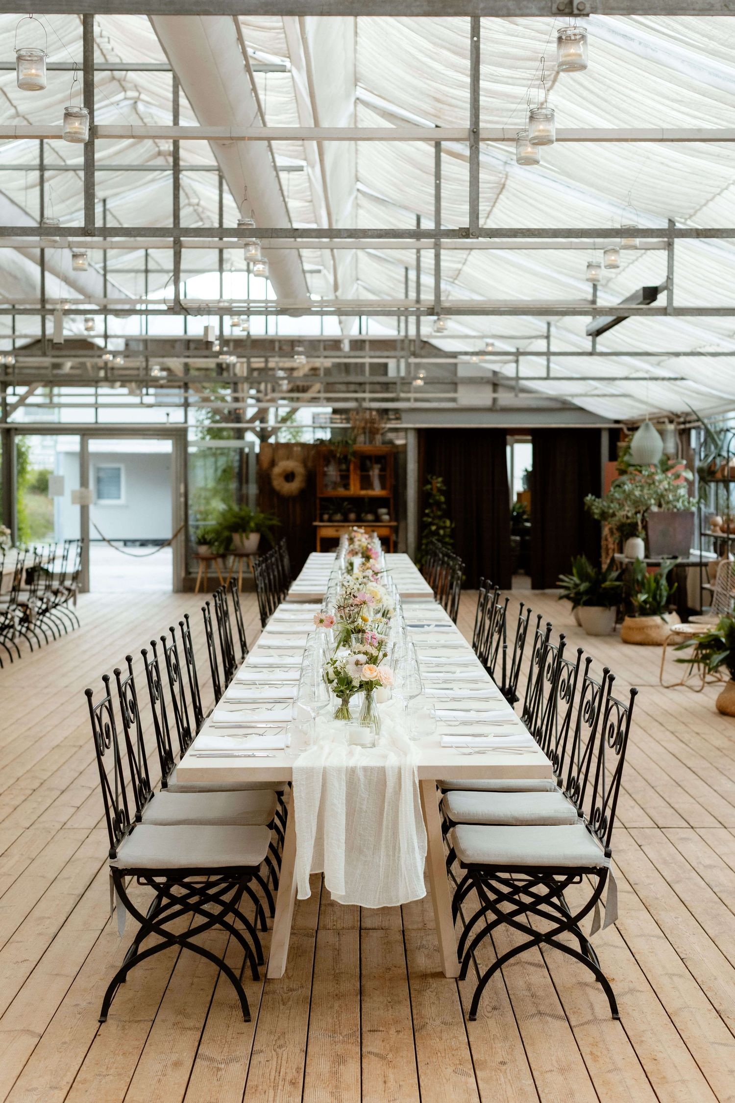 Elegant greenhouse wedding reception with long white-draped table and black iron chairs.