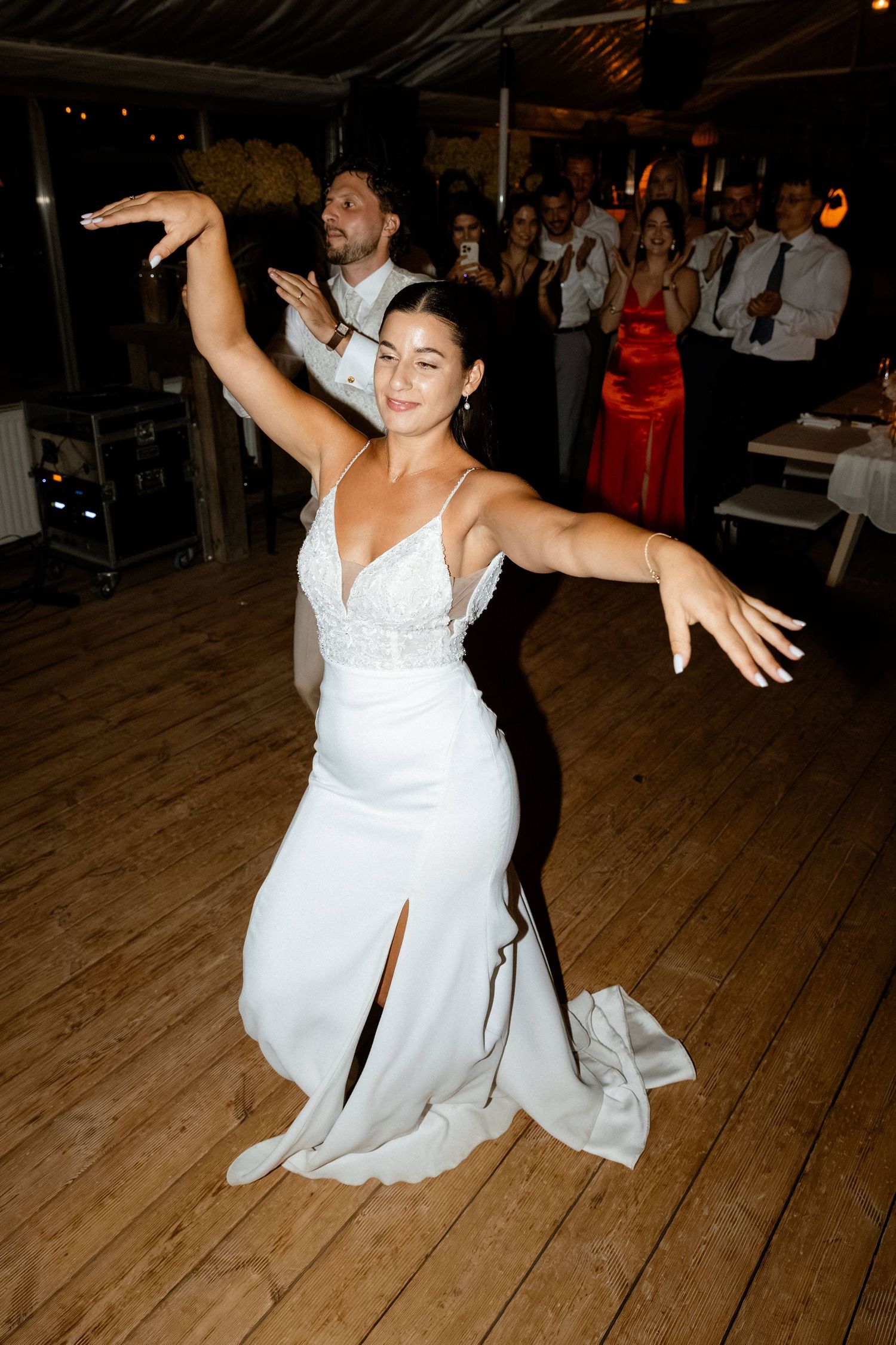 Bride in elegant white slit gown dancing with arms outstretched on wooden dance floor at wedding.