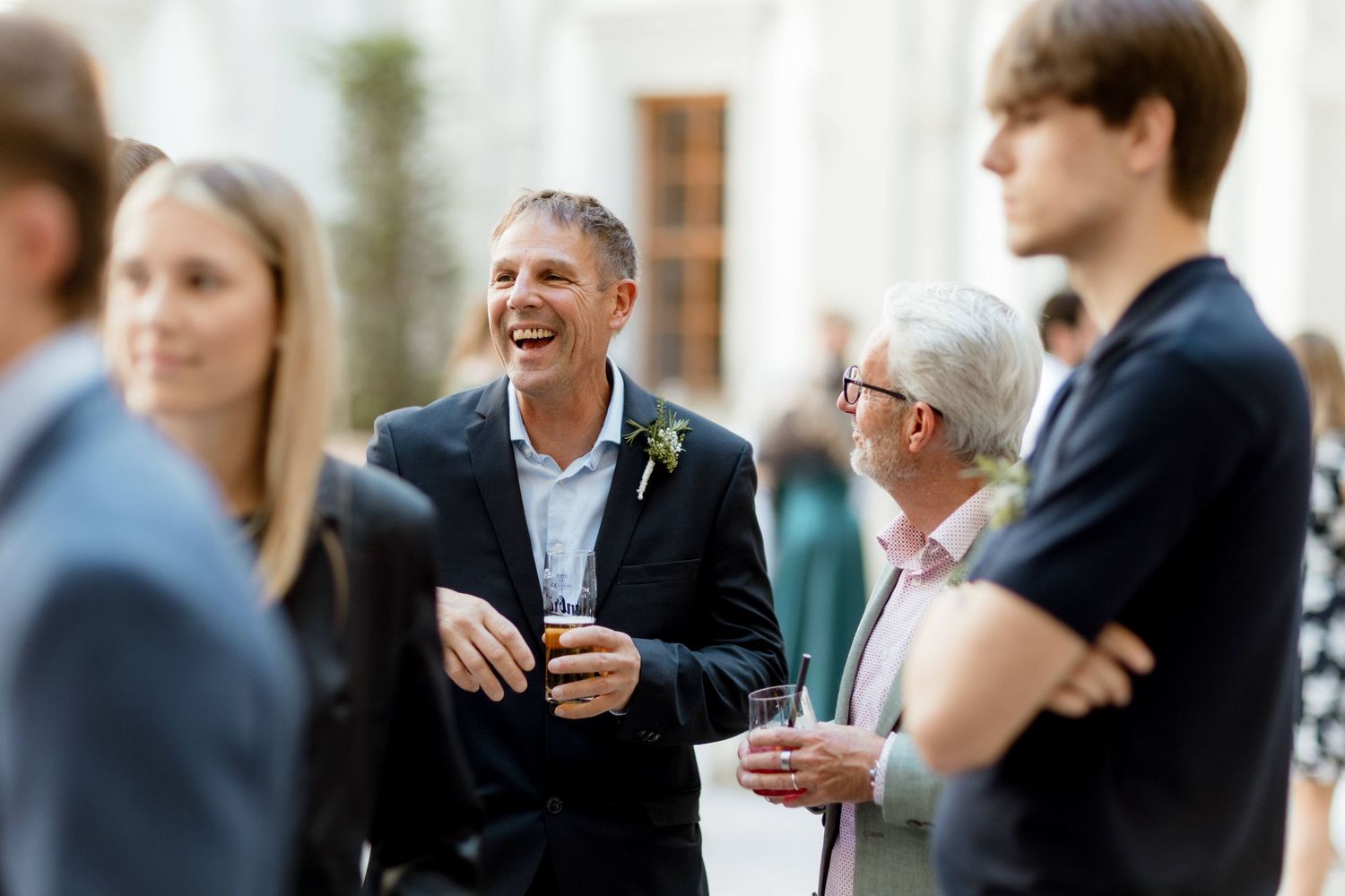 Laughing man in dark blazer with boutonniere socializing with guests at outdoor wedding reception.