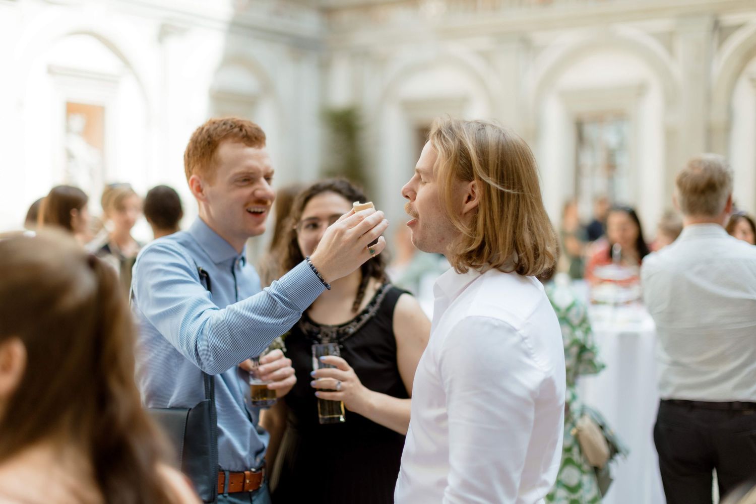 Red-haired man playfully feeding a friend at a lively wedding reception gathering indoors.
