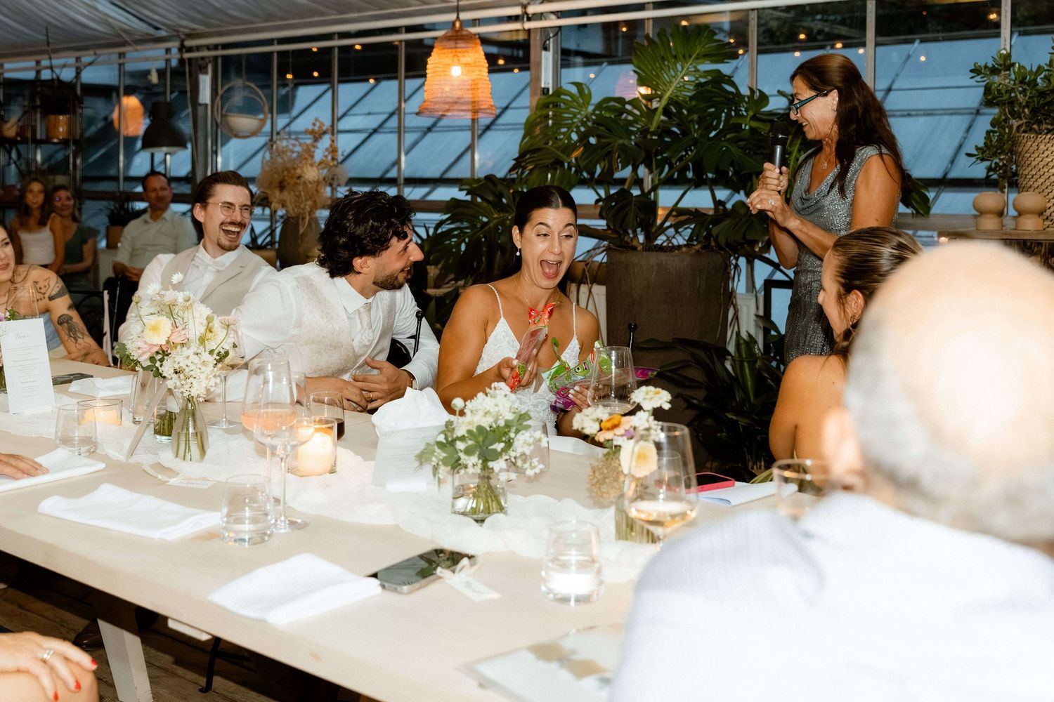 Joyful wedding guests laughing together at elegant reception table with floral centerpieces.