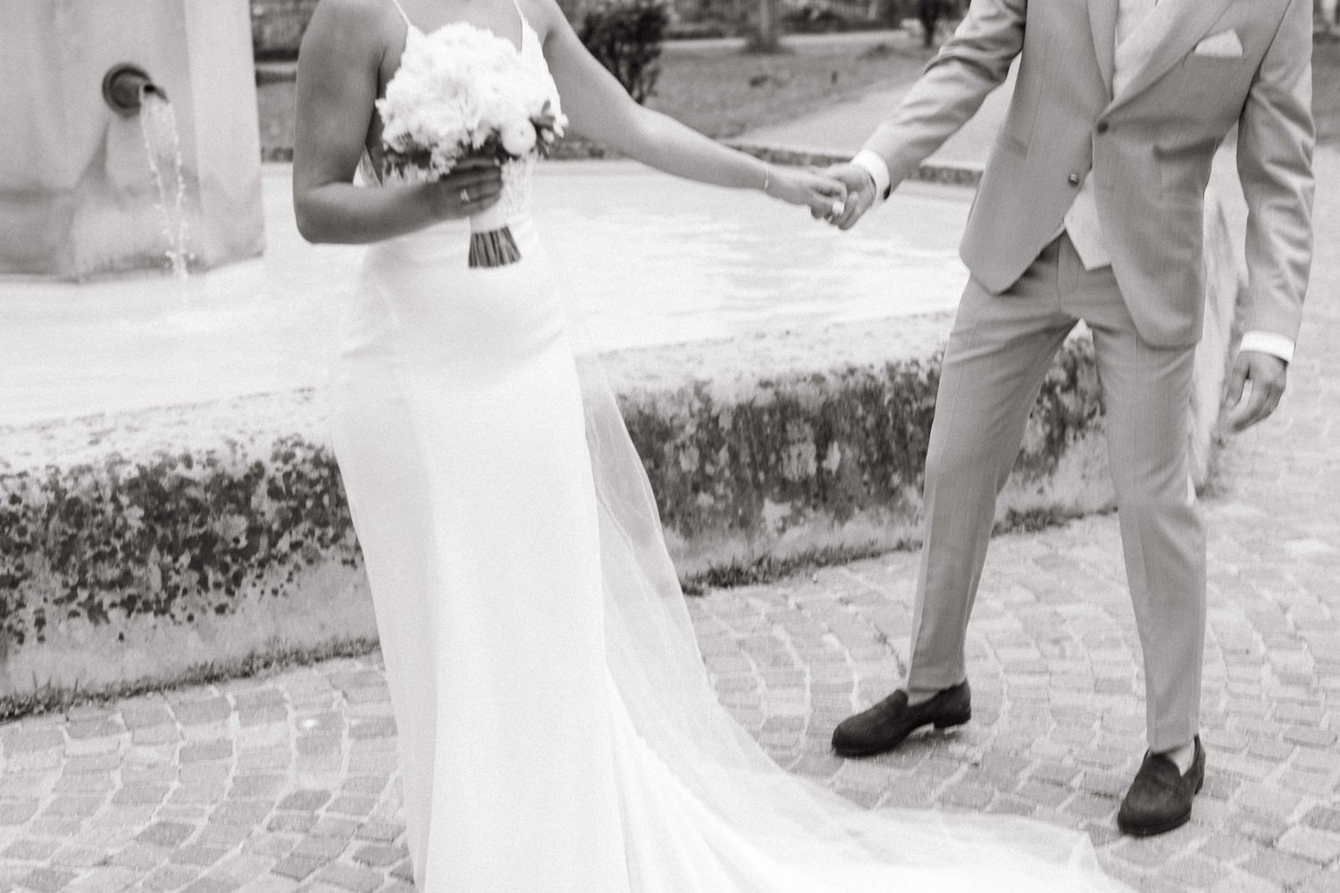 Black and white photo of bride and groom holding hands near fountain on wedding day.