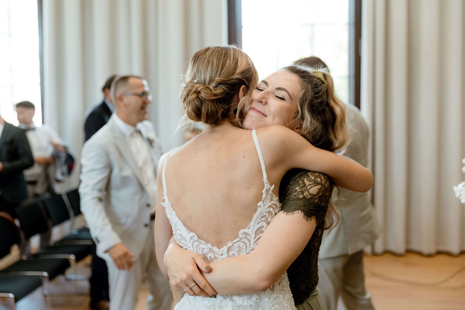 Bride in lace wedding gown sharing a heartfelt hug with a female guest at wedding reception.