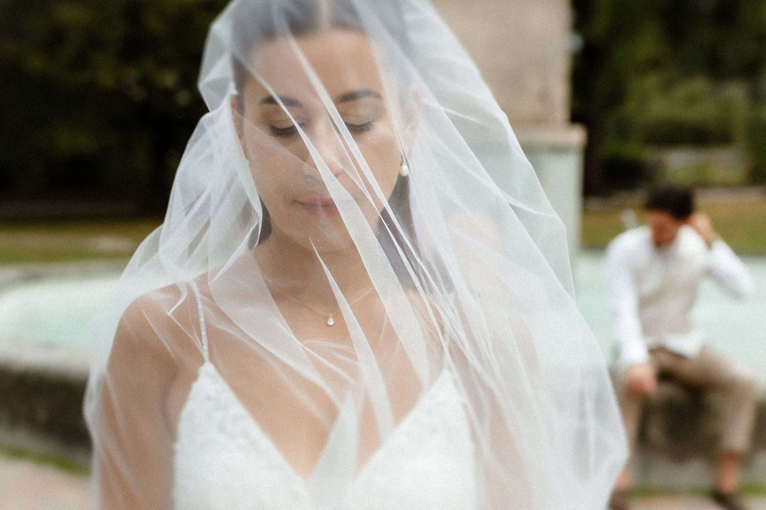 Bride in white wedding dress covered by flowing veil with fountain in background.