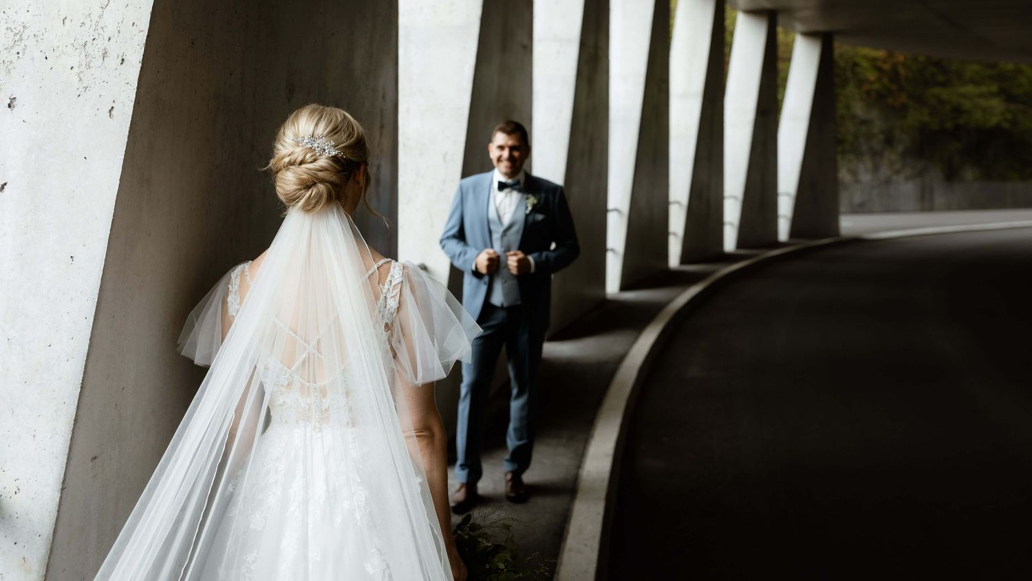 Bride in white gown and veil walks toward groom in blue suit under concrete bridge.