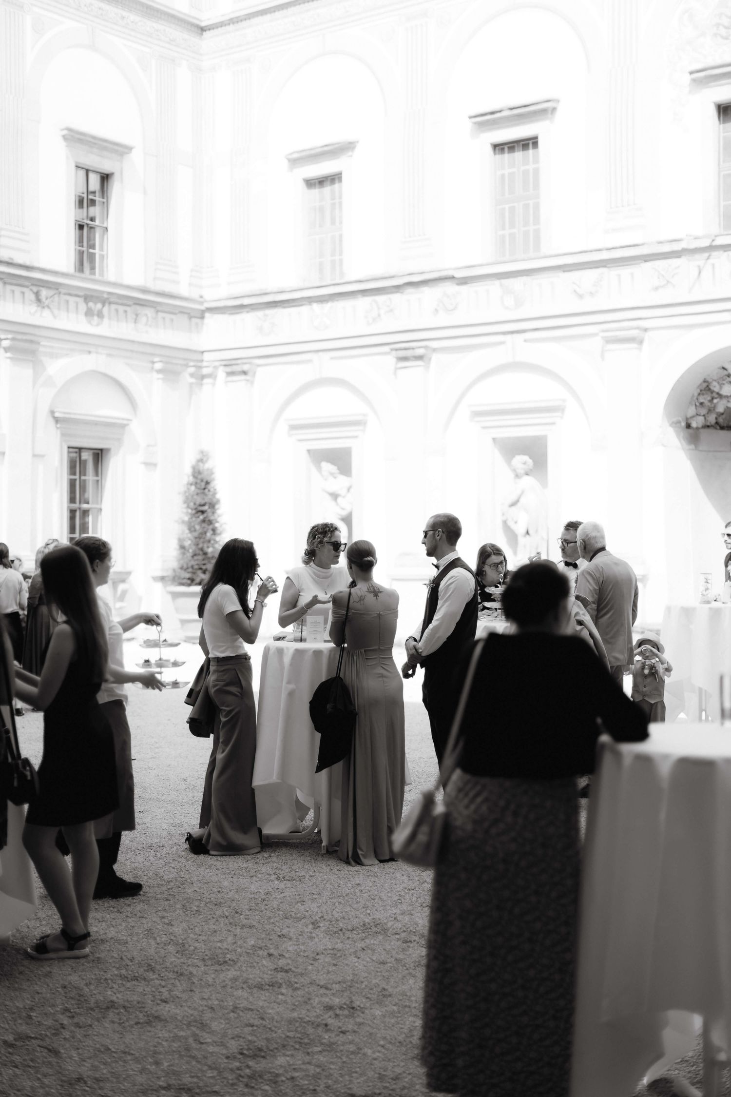 Black and white photo of wedding guests socializing in an elegant courtyard with classical statues and arches.