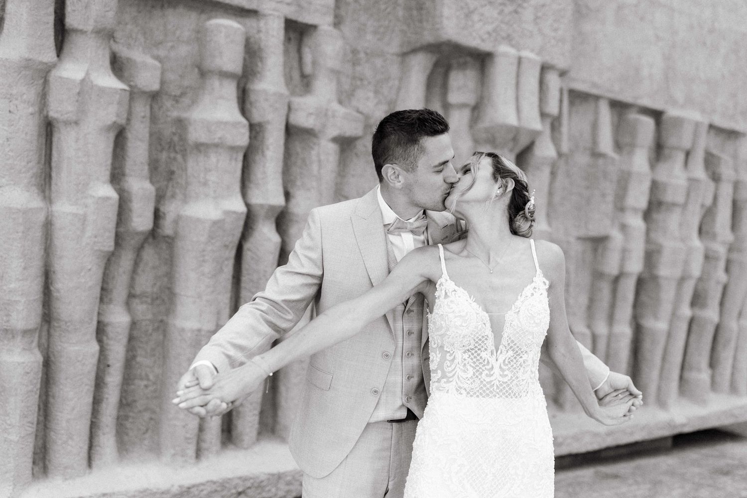 Black and white photo of bride and groom sharing a romantic kiss near ornate stone wall on wedding day.
